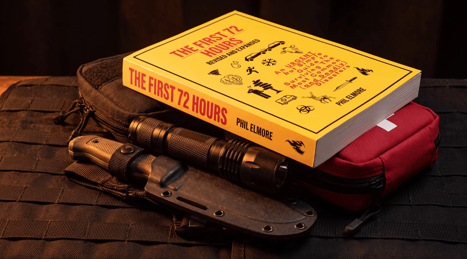 The First 72 Hours by Phil Elmore, revised and expanded edition, sitting on a tactical pack next to a fixed blade knife, a flashlight and a red medical pouch