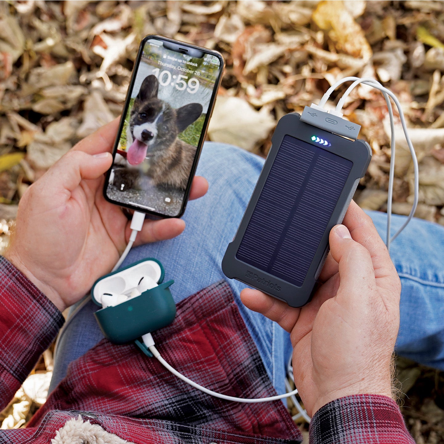 A solar power bank with a panel on the front being held outdoors while charging a phone and a pair of earbuds from two of its USB ports