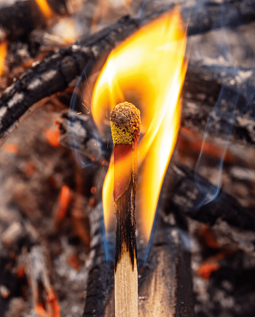 Close view of a lit stormproof match burning with a tall flame above a bed of embers, showing the thick coated head